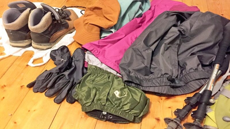 富士登山の初心者に必要な持ち物一式。登山靴、レインウェア、帽子、手袋、ゲイター、ストックなどが床に並べられている。