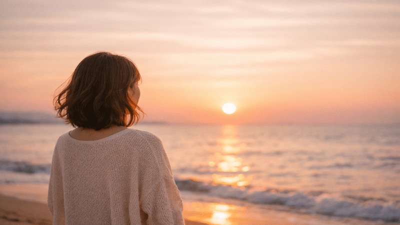 夕焼けの海を見つめる女性の後ろ姿。静かな時間の中で自分と向き合うイメージ